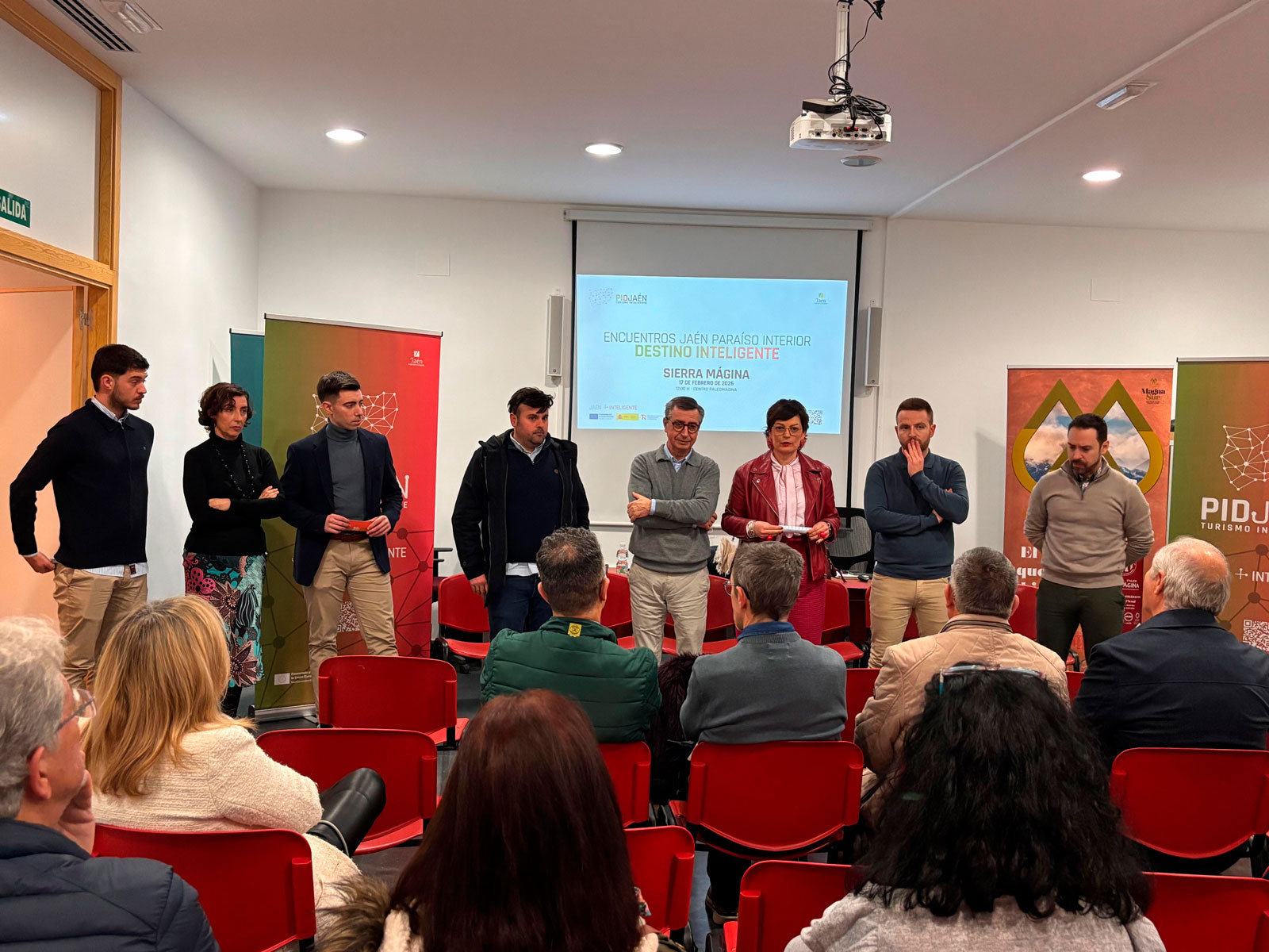 Encuentro comercial PID Jaén: grupo de personas de pie y sentadas en sillas rojas durante una presentación, con proyector y paneles informativos de fondo en sala de eventos