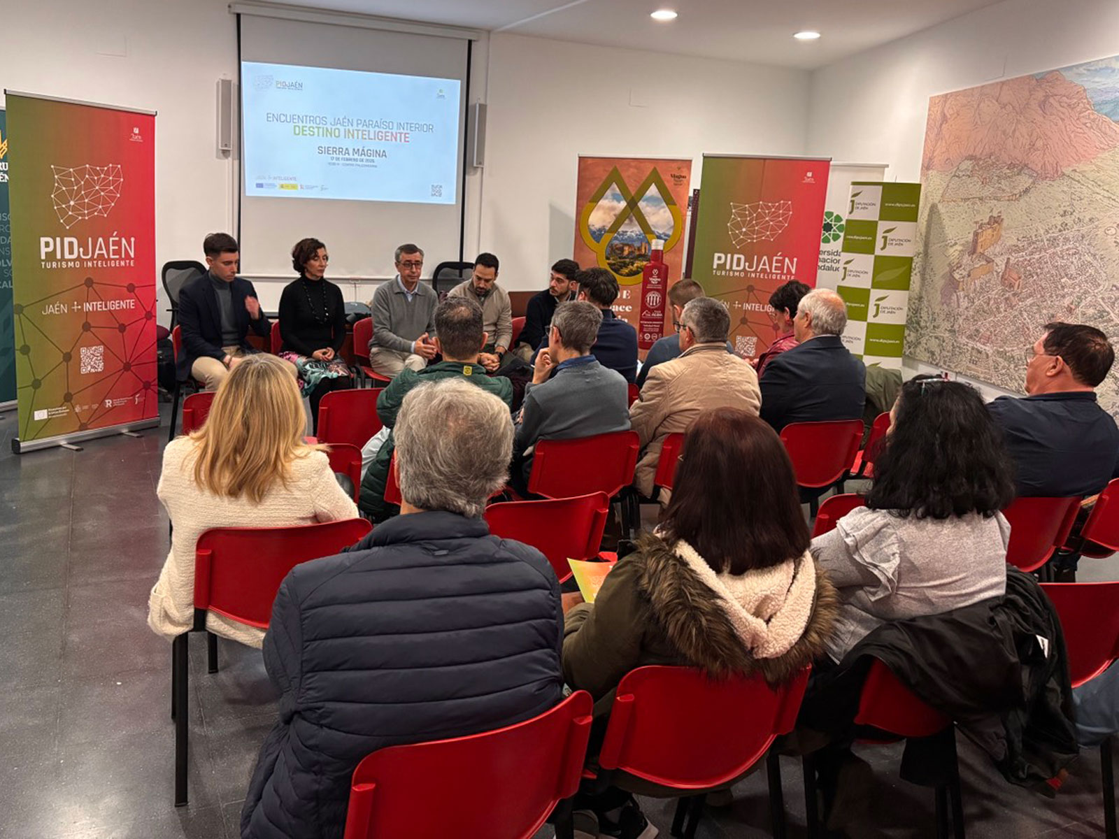 Panel de expertos y público participando en evento del Proyecto PID Jaén sobre Turismo Inteligente, con banners de la Diputación y carteles informativos en la sala