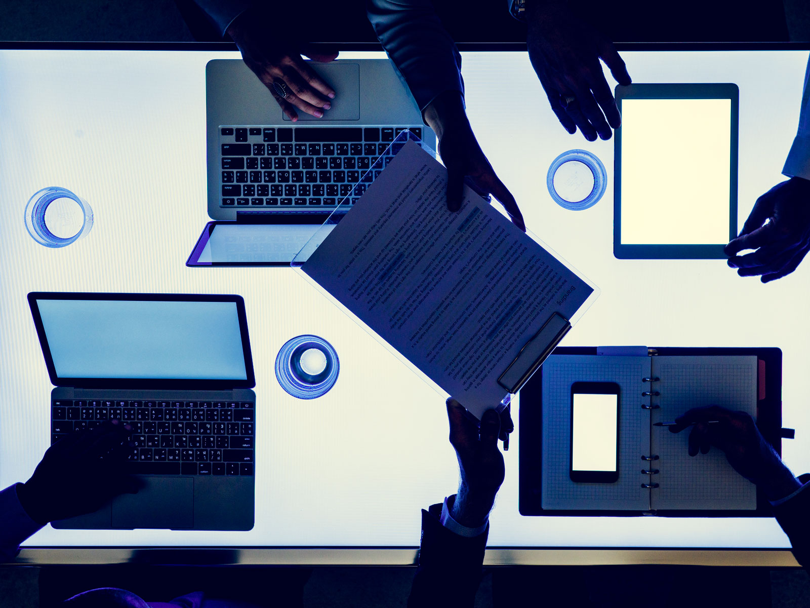 Equipo colaborativo trabajando con diversos dispositivos digitales incluyendo laptop, tablet y portapapeles digitales en ambiente de trabajo moderno con elementos de interfaz gráfica azul